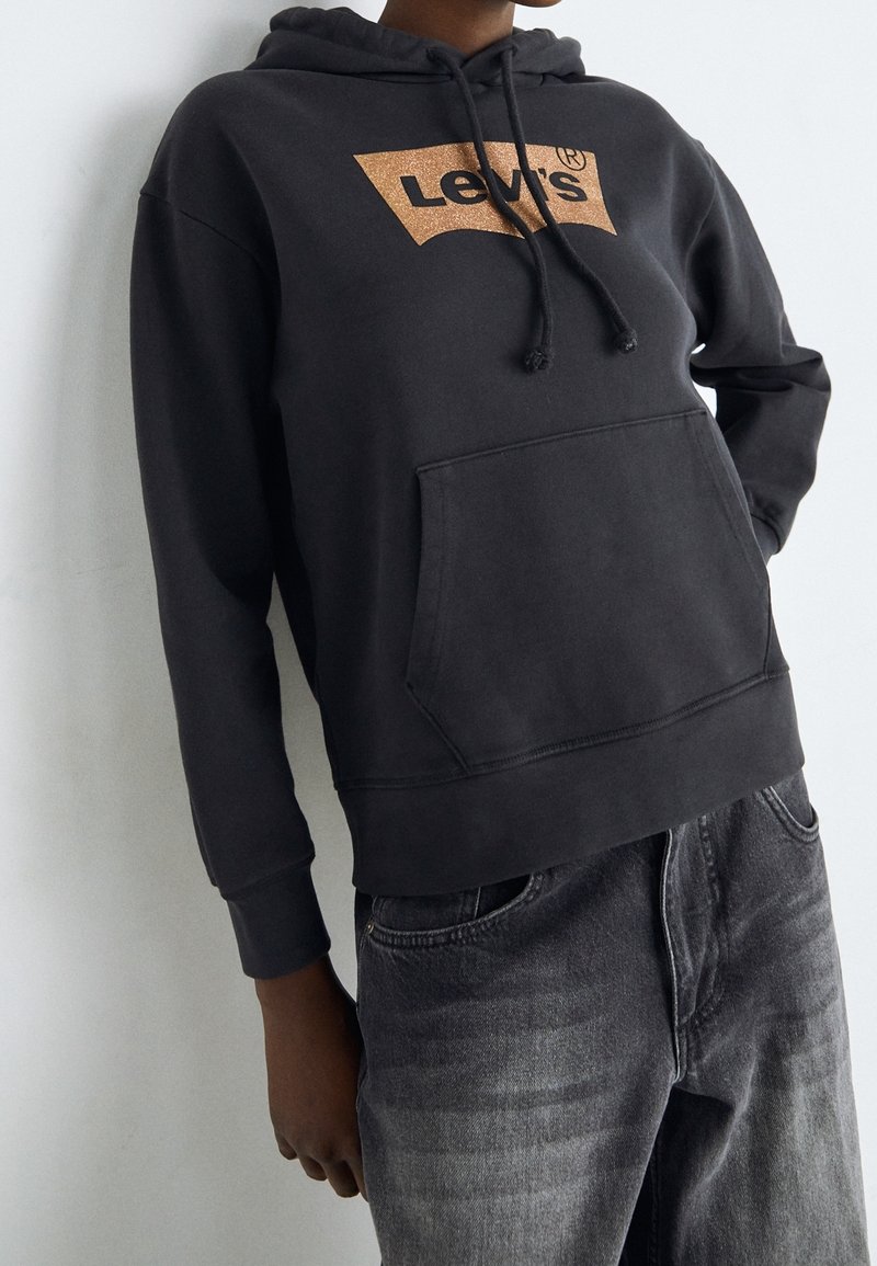 Personne portant un sweat à capuche noir Levi's avec une poche avant et un jean noir délavé, debout devant un fond clair uni.