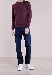 Sudadera de cuello redondo en color burdeos con el logo en relieve "BOSS", combinada con jeans azul oscuro y zapatillas blancas. Textura suave, ajuste casual.
