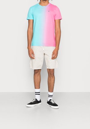 Hombre que lleva una camiseta mitad azul, mitad rosa, pantalones cortos blancos con rayas verticales negras, calcetines blancos con rayas negras y zapatillas negras.