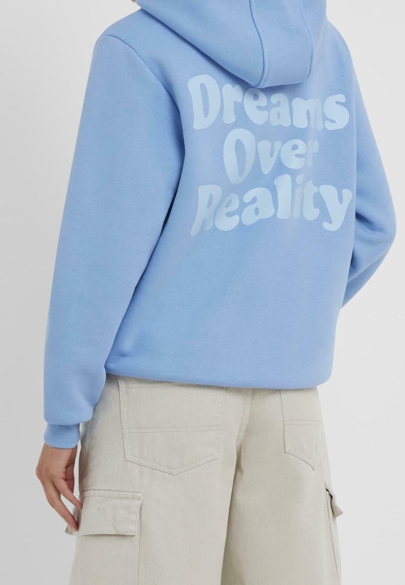 Personne portant un sweat à capuche bleu clair avec le texte "Dreams Over Reality" dans le dos et un pantalon cargo beige, debout avec la main près de la poche.