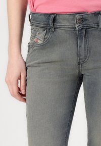 Grå denimjeans med slim fit, slitte detaljer på lommene og et synlig merketagg på siden. Nærbilde av linning.
