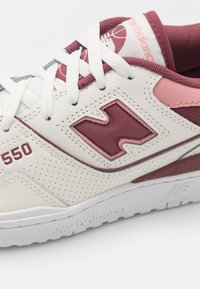 Baskets New Balance 550 dotées d'une tige en cuir crème avec des accents bordeaux et des détails perforés, des lacets blancs et une semelle en caoutchouc texturée.