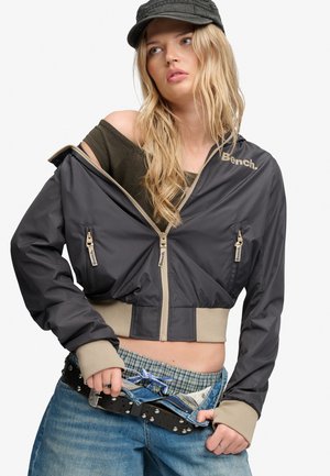 Junge Frau mit langen blonden Haaren trägt eine schwarze Kappe, eine schwarze kurze Jacke, ein grünes kariertes Hemd und blaue Jeans mit einem Nietengürtel.