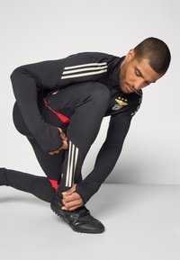 Zwart sportoutfit met rode accenten, met drie witte strepen op de mouwen en ritsen op de broek. Zwarte schoenen maken de look compleet.