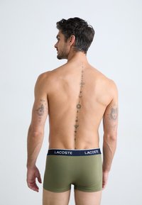 Olijfgroene herenboxershorts met een marineblauwe tailleband met het "Lacoste" logo. Huid zichtbaar; ingewikkelde tatoeages op de rug en armen.