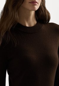 Brun stickad tröja med ribbad, hög krage. Mjuk textur och åtsittande design som visar en enkel och modern look.