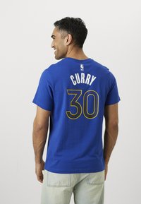 Tricou din bumbac de culoare albastru regal, cu guler rotund, având inscripția "CURRY" și numărul "30" imprimate în alb și galben pe spate, logo-ul NBA deasupra.