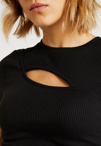 Kvinna i en svart ribbad topp med en unik asymmetrisk utskärning över den övre bröstkorgen och axeln.
