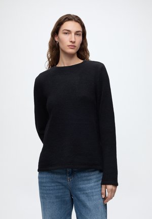 Femme portant un pull noir à manches longues et un jean bleu, debout devant un fond blanc uni.