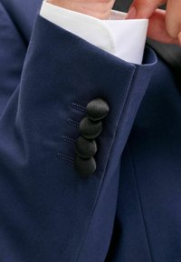 Primo piano di una persona che sistema il polsino di una giacca da completo blu navy con quattro bottoni rivestiti in tessuto nero e il polsino di una camicia bianca elegante.