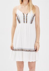 Weißes ärmelloses Kleid mit V-Ausschnitt, verziert mit bunten Stickereien entlang des Oberteils und einem voluminösen Saum. Leichte Stoffqualität.
