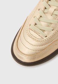 Χρυσό μεταλλικό αθλητικό παπούτσι με λείο υφασμένο επάνω μέρος, κρεμ κορδόνια και καφέ ελαστική σόλα. Διαθέτει διακοσμητικές ραφές κατά μήκος των πλευρών.