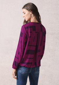 Blusa viola con un motivo geometrico rosa, maniche lunghe, scollatura rotonda e polsini elasticizzati, abbinata a jeans di denim.