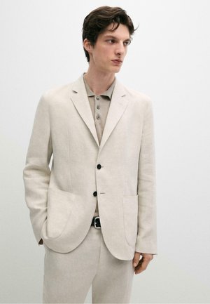 Giovane uomo che indossa un abito beige con bottoni neri e una polo marrone chiaro, in piedi con una mano in tasca, guarda verso destra.