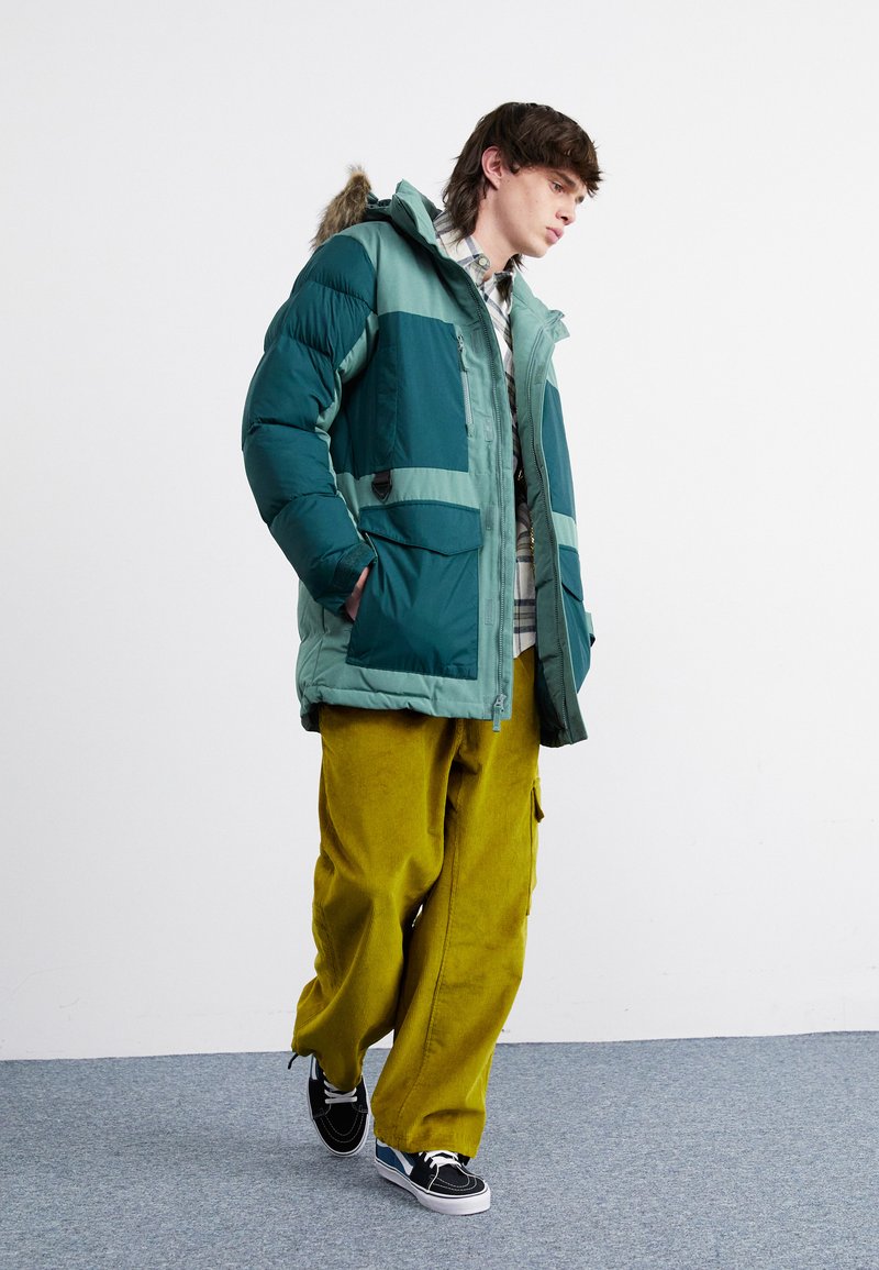 Parka matelassée en teal et vert avec capuche en fourrure, dotée de grandes poches et d'une fermeture éclair à l'avant, associée à un pantalon en velours côtelé jaune et des baskets noires.