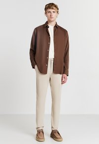 Un jeune homme portant une chemise marron à boutons sur un t-shirt blanc, un pantalon beige et des chaussures en suède marron, se tenant devant un fond blanc uni.