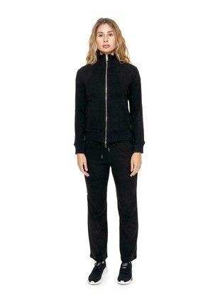 Une femme porte une veste noire zippée, un pantalon assorti et des baskets sur fond blanc.