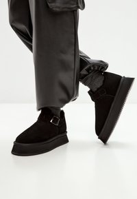 Person iført sorte ruskind ankelstøvler med spænderemme, grå sokker og løse sorte læderbukser mod en hvid baggrund.