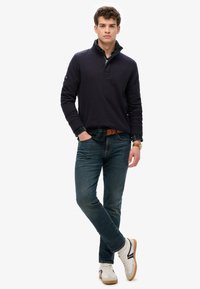 Marineblauwe pullover met hoge kraag, gedragen over een geblokt shirt; gecombineerd met donkere jeans en witte sneakers; gladde textuur, casual stijl.