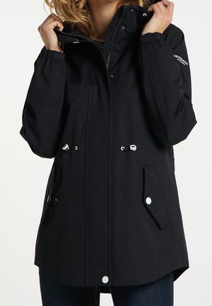 Kvinde iført sort hættetrøje med snore, lommer med trykknapper og lynlås, der holder kraven med begge hænder.