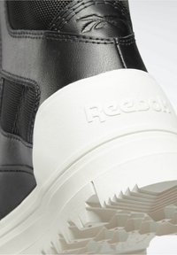 Black leather sneaker with textured mesh upper and white rubber sole; features Reebok logo embossed on heel and zigzag tread pattern.