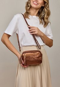Metalická bronzová crossbody taška se dvěma zipovými oddíly a třásňovými akcenty. Obsahuje odnímatelný popruh a hladký povrch.