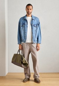 Chaqueta de mezclilla en azul claro, camiseta blanca, pantalones beige, bolsa de lona verde con detalles en marrón y zapatos de ante grises.