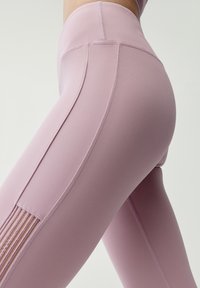 Jasnoróżowe legginsy sportowe o gładkiej teksturze, z wysokim stanem i bocznym panelem z pionowymi wycięciami. Widoczne detale przeszyć.