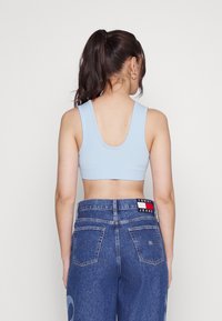 Světle modrý žebrovaný crop top s širokými ramínky, kombinovaný s tmavě modrými džíny s vysokým pasem, které mají viditelnou štítkovou etiketu na pásku.