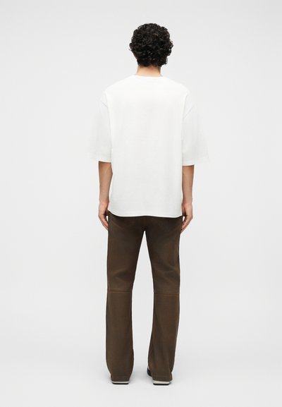 Göndör fekete hajú személy háttal áll, laza, fehér rövid ujjú inget és egyenes barna nadrágot visel, egyszerű háttér előtt.