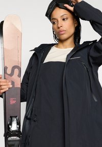 Μαύρο μπουφάν χιονιού με κουκούλα, υφή ύφασμα και τσέπες με φερμοάρ; ανοιχτόχρωμο τοπ; κρατάει ροζ σκι με μαύρους δέντρες.