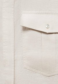 Beige linen shirt featuring a patch pocket with a contrasting button, textured fabric, and subtle stitching details.