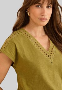 Moteris su ilgais rudais plaukais, dėvinti auksines auskarų žiedus ir alyvuogių spalvos palaidinę su akių nėriniais apykaklėje ir rankovėse.