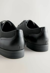 O pereche de pantofi de dress din piele neagră și material textil, cu detalii cusute, văzuți din spate pe un fundal deschis și neutru.