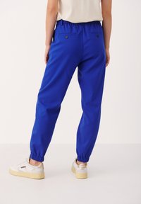 Blå joggingsbyxor med elastisk midja, elastiska muddar och två bakfickor, i kombination med vita sneakers. Slät tygtextur.