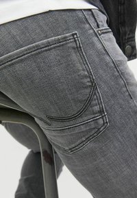 Jeans din denim gri, prezentând un buzunar pe spate cu un design de cusătură curbat. Țesătura pare ușor uzată, având o textură netedă.