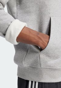 Ljusgrå sweatshirt med mjuka vita fleeceärmar, har en framficka och en avslappnad passform. Detaljerad sömnad synlig nära fållen.