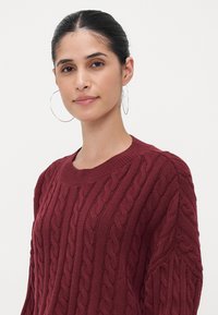 Cable-knit burgundy sweater with a wide crew neckline, featuring a textured pattern and loose-fitting sleeves. Soft, ribbed fabric.