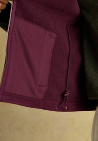 Fialová fleece podšívka bundy, ktorá má veľkú kapsu, detaily sťahovacej šnúrky a kontrastnú tmavú tkaninu na vonkajšej strane.