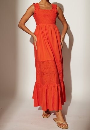 Frau trägt ein ärmelloses oranges Maxikleid mit gerafften Trägern und Saum, mit dekorativen perforierten Mustern, steht mit den Händen in der Hüfte.