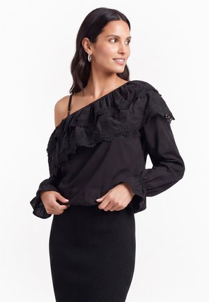 Donna con lunghi capelli scuri indossa una camicetta nera monospalla con volant e una gonna nera a coste, guarda verso destra con un leggero sorriso.