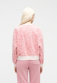 Γυναίκα με μακριά ξανθά μαλλιά που φορεί ροζ πουλόβερ με λεοπάρ σχέδιο και ροζ παντελόνι, εμφανιζόμενη από πίσω με φόντο απλό.