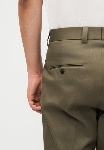 Κοντινή λήψη ενός ατόμου που φοράει παντελόνι ελιάς με κουμπωμένη πίσω τσέπη και άσπρο πουκάμισο μπαλαρισμένο μέσα.