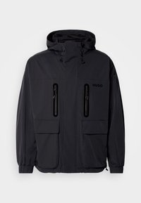 Black waterproof jacket with a hood, two zippered chest pockets, and elastic cuffs. Features minimalistic design and "HUGO" branding.