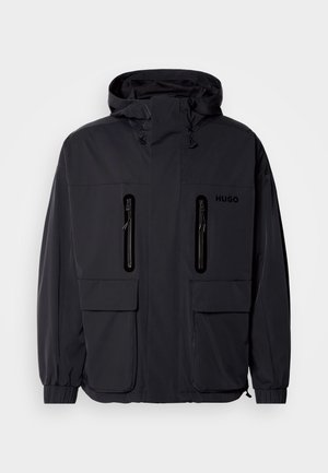 Veste imperméable noire avec capuche, deux poches poitrine zippées et poignets élastiques. Présente un design minimaliste et un logo "HUGO".