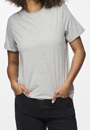 Osoba ubrana w jednolity jasnoszary t-shirt z krótkim rękawem i czarne spodnie, z rękami w kieszeniach, wykadrowana powyżej ramion.