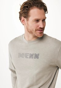 Felpa grigia chiara realizzata in tessuto morbido, con un logo testurizzato "MEXX" in grigio scuro sul petto. Collo rotondo e maniche lunghe.