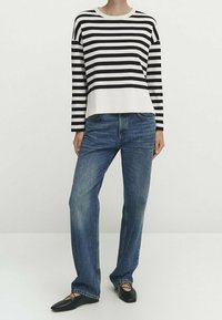 Henkilö, joka pukeutuu musta-valkoiseen vaakaraidalliseen pitkähiharaiseen paitaan, sinisiin suoriin farkkuihin ja mustiin nilkkatuilla ballerinatossuihin.