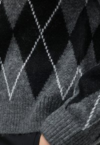 Gros plan d'un pull en laine gris et noir à motif losanges avec des poignets côtelés sur le bras d'une personne.