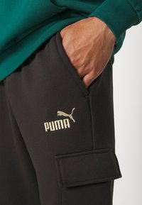 Černé bavlněné tepláky s cargo kapsou, zlatým logem Puma a rukou umístěnou v kapse. Zelená mikina viditelná nahoře.